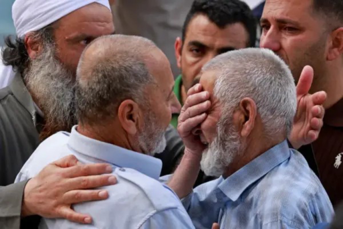 Two older men in light shirts share a comforting embrace as others surround them in a tense moment.