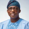 Portrait of a smiling man wearing a light blue traditional outfit with a patterned headwrap and round glasses.