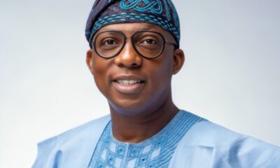 Portrait of a smiling man wearing a light blue traditional outfit with a patterned headwrap and round glasses.
