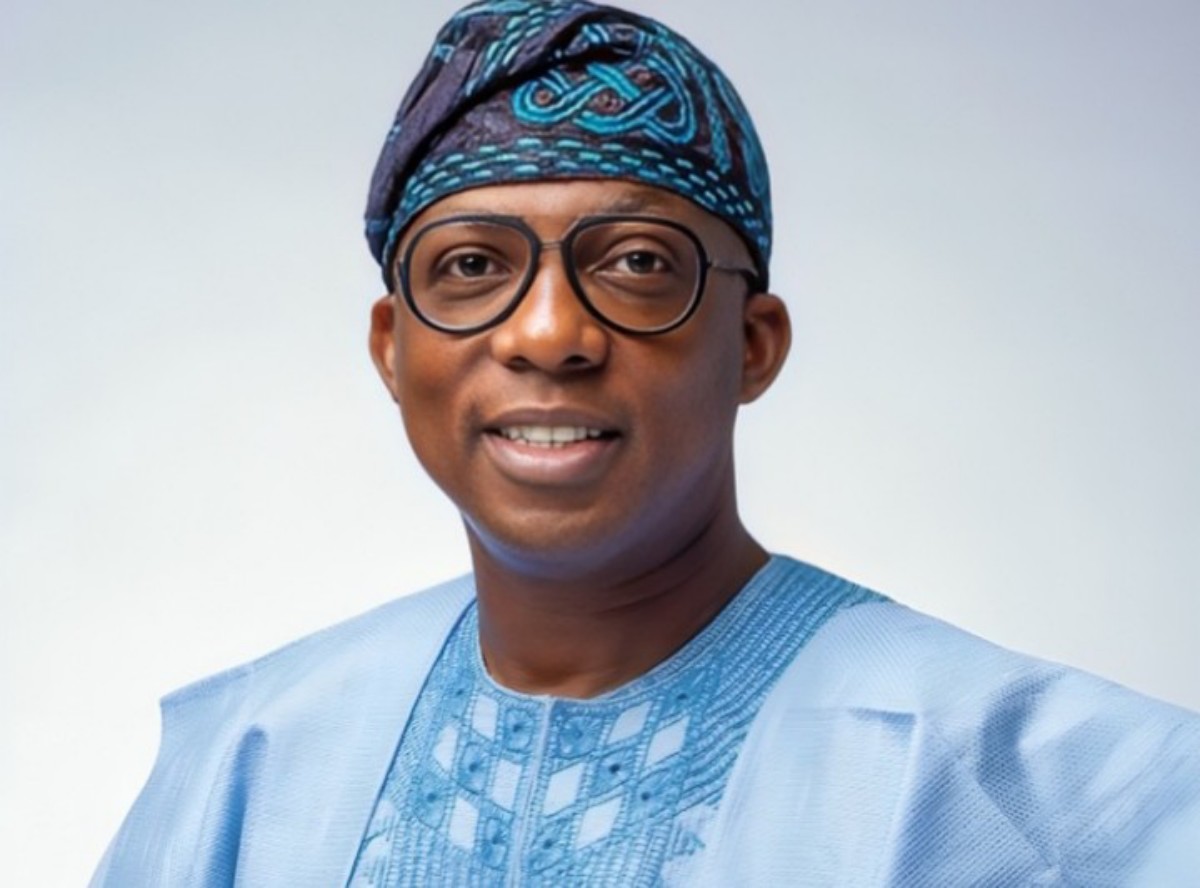 Portrait of a smiling man wearing a light blue traditional outfit with a patterned headwrap and round glasses.