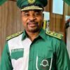 Smiling man in a green military-style uniform and cap, standing indoors with a friendly expression (name badge reads MC Oluomo).