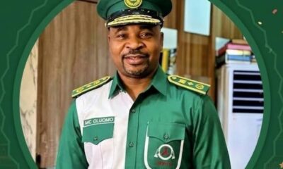 Smiling man in a green military-style uniform and cap, standing indoors with a friendly expression (name badge reads MC Oluomo).
