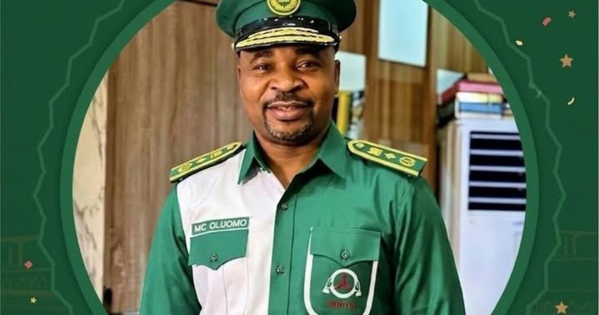 Smiling man in a green military-style uniform and cap, standing indoors with a friendly expression (name badge reads MC Oluomo).