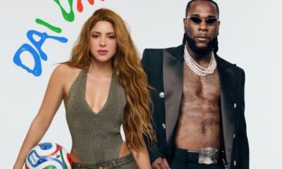 Woman with long red hair in an olive-green halter top holding a soccer ball beside a man in a black blazer with chains and sunglasses.