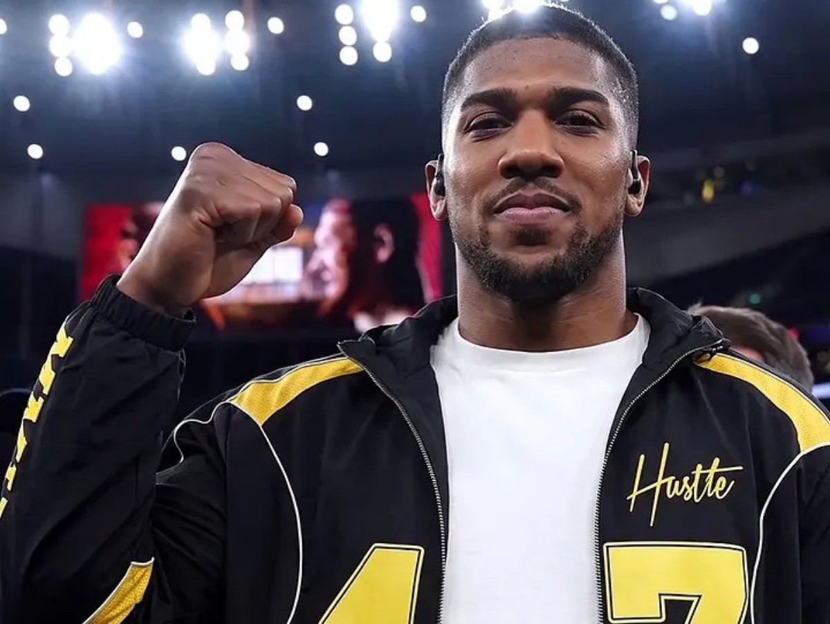 Male athlete raises a clenched fist in victory, wearing a black and yellow jacket with 'Hustle' on the chest in a bright arena.