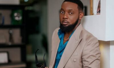 Man with a beard wearing a tan blazer and teal shirt, standing indoors and holding sunglasses in one hand.