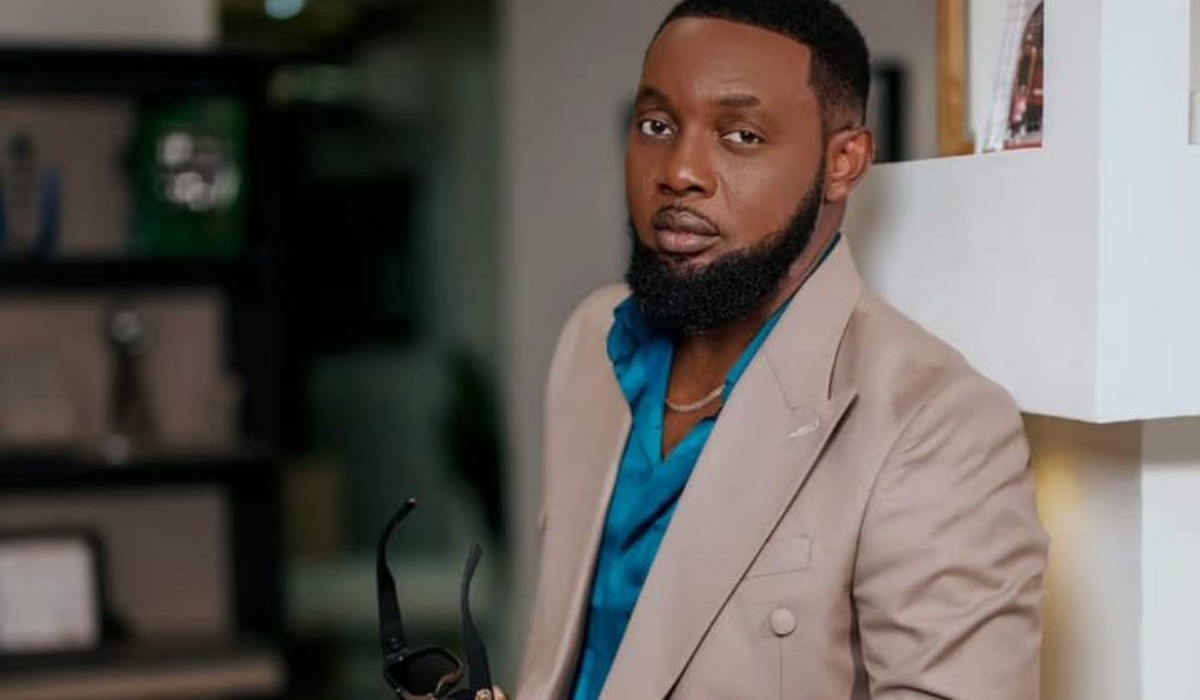 Man with a beard wearing a tan blazer and teal shirt, standing indoors and holding sunglasses in one hand.