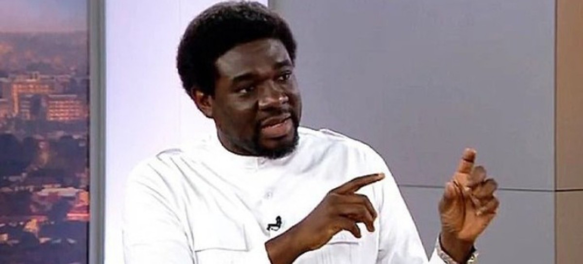 Man in a white shirt speaks in a studio, gesturing with his hands as he makes a point.