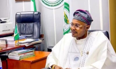 Man in a white traditional outfit and head wrap sits at a desk in an office, with flags and stacks of papers nearby.
