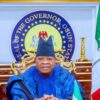 Official portrait of Osun State governor seated at a ceremonial desk with flags and the state seal behind him, wearing blue traditional attire and a cap.