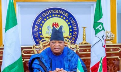 Official portrait of Osun State governor seated at a ceremonial desk with flags and the state seal behind him, wearing blue traditional attire and a cap.