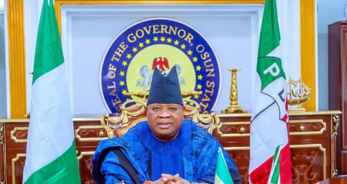 Official portrait of Osun State governor seated at a ceremonial desk with flags and the state seal behind him, wearing blue traditional attire and a cap.
