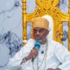 Man wearing a light-colored traditional outfit and orange turban speaks into a blue microphone while seated on a gold ornate throne.