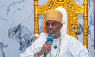 Man wearing a light-colored traditional outfit and orange turban speaks into a blue microphone while seated on a gold ornate throne.