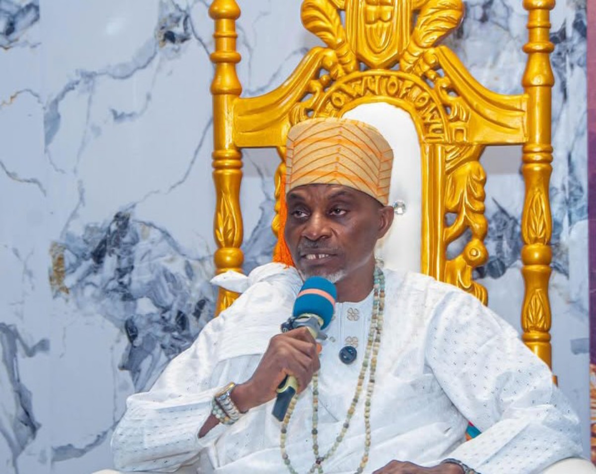 Man wearing a light-colored traditional outfit and orange turban speaks into a blue microphone while seated on a gold ornate throne.