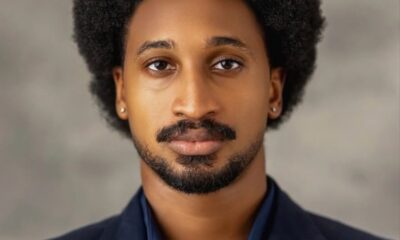 Portrait of a man with a large Afro hairstyle wearing a navy blazer and dark shirt, looking at the camera.