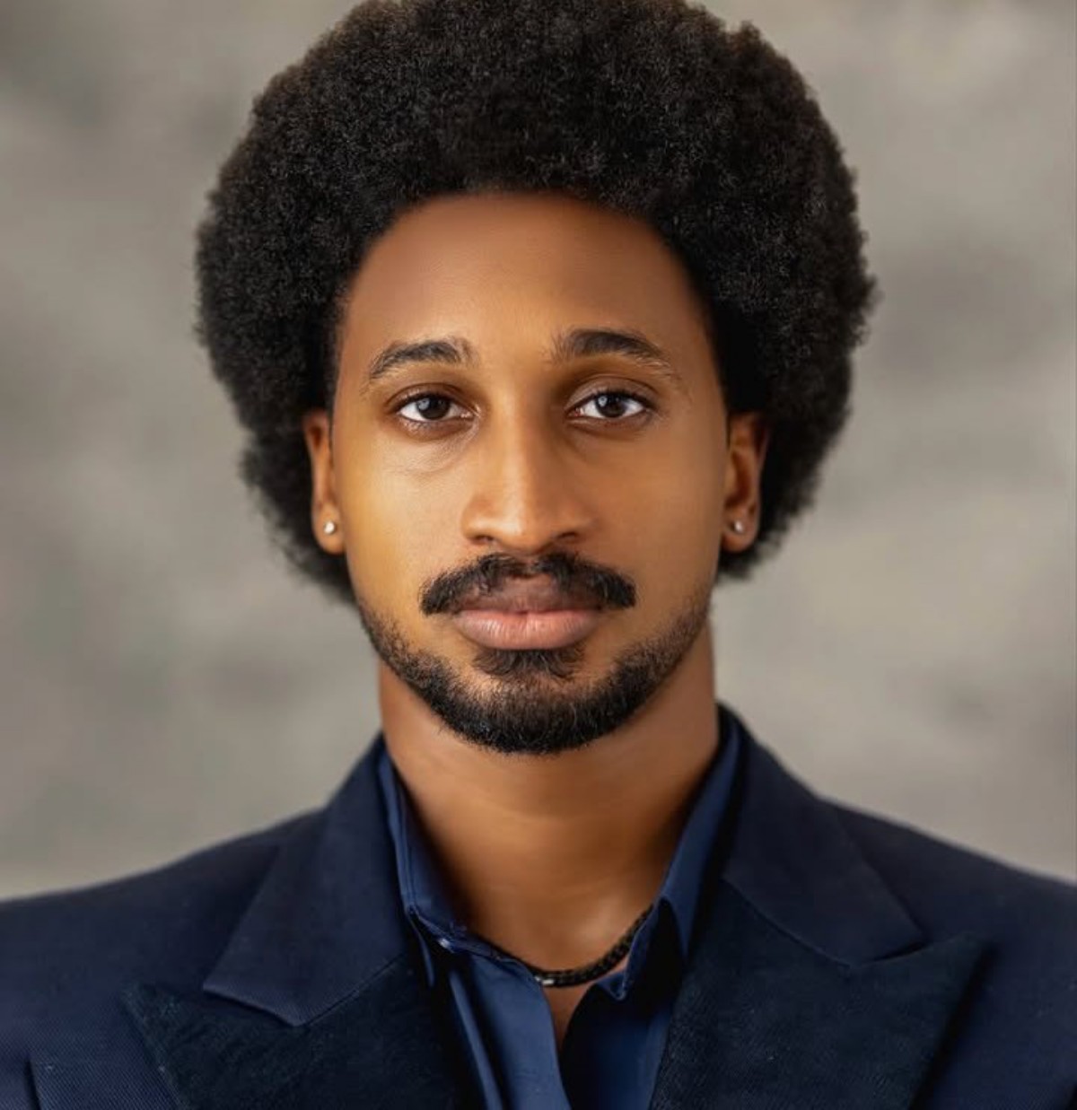 Portrait of a man with a large Afro hairstyle wearing a navy blazer and dark shirt, looking at the camera.