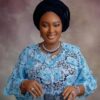 Portrait of a smiling woman wearing a light blue lace dress and a black headwrap, with silver jewelry against a muted backdrop.