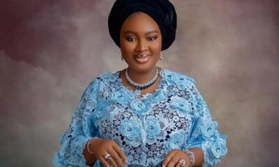 Portrait of a smiling woman wearing a light blue lace dress and a black headwrap, with silver jewelry against a muted backdrop.