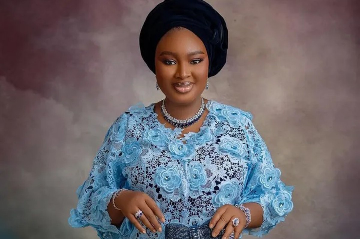Portrait of a smiling woman wearing a light blue lace dress and a black headwrap, with silver jewelry against a muted backdrop.