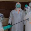 Four healthcare workers in full PPE (hazmat suits, face shields) perform disinfection with a spray wand and yellow tank nearby, indoors.
