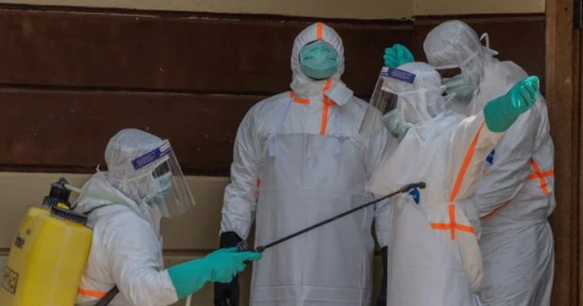 Four healthcare workers in full PPE (hazmat suits, face shields) perform disinfection with a spray wand and yellow tank nearby, indoors.