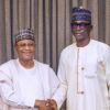 Two Nigerian men in white traditional outfits shake hands and smile in front of a patterned curtain.