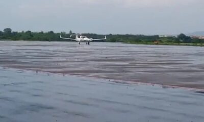 White twin-engine amphibious aircraft on a water-covered airstrip with a distant green shoreline behind it of trees and fields.