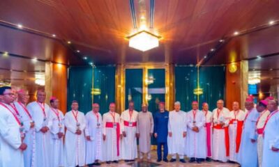 Group of clergymen in white robes with red sashes posing for a formal group photo in a gilded hall.