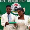 Wedding couple in white attire stand before a green Nigerian banner; groom holds a green scroll while the bride carries a white bouquet of baby's breath.