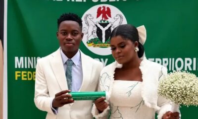 Wedding couple in white attire stand before a green Nigerian banner; groom holds a green scroll while the bride carries a white bouquet of baby's breath.