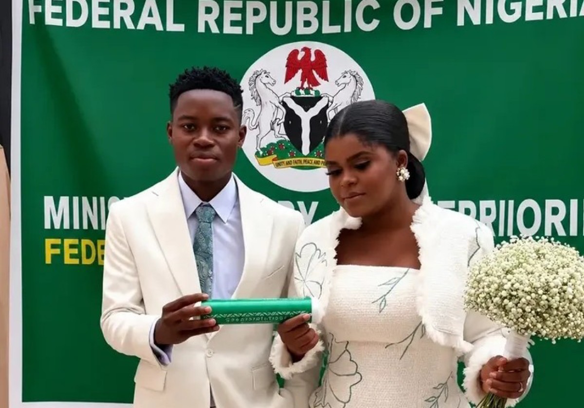 Wedding couple in white attire stand before a green Nigerian banner; groom holds a green scroll while the bride carries a white bouquet of baby's breath.