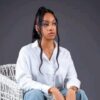 Woman seated in a white wicker chair wearing a white button-down shirt and light blue jeans against a gray studio backdrop.