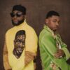 Two men pose back-to-back in stylish outfits: one in a yellow turtleneck with a graphic face, wearing a black cap and sunglasses, the other in a bright green embellished jacket with a scarf and jewelry.