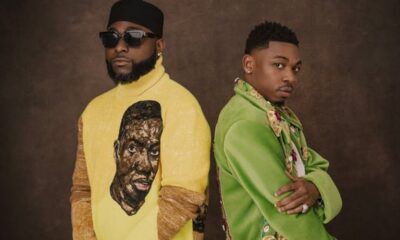 Two men pose back-to-back in stylish outfits: one in a yellow turtleneck with a graphic face, wearing a black cap and sunglasses, the other in a bright green embellished jacket with a scarf and jewelry.