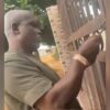 Man with a shaved head in a khaki shirt reaches to open a metal gate with bars.
