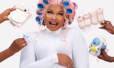 Pregnant woman smiling with pink-blue hair rollers, surrounded by hands offering baby items like wipes, socks, and plush toy.