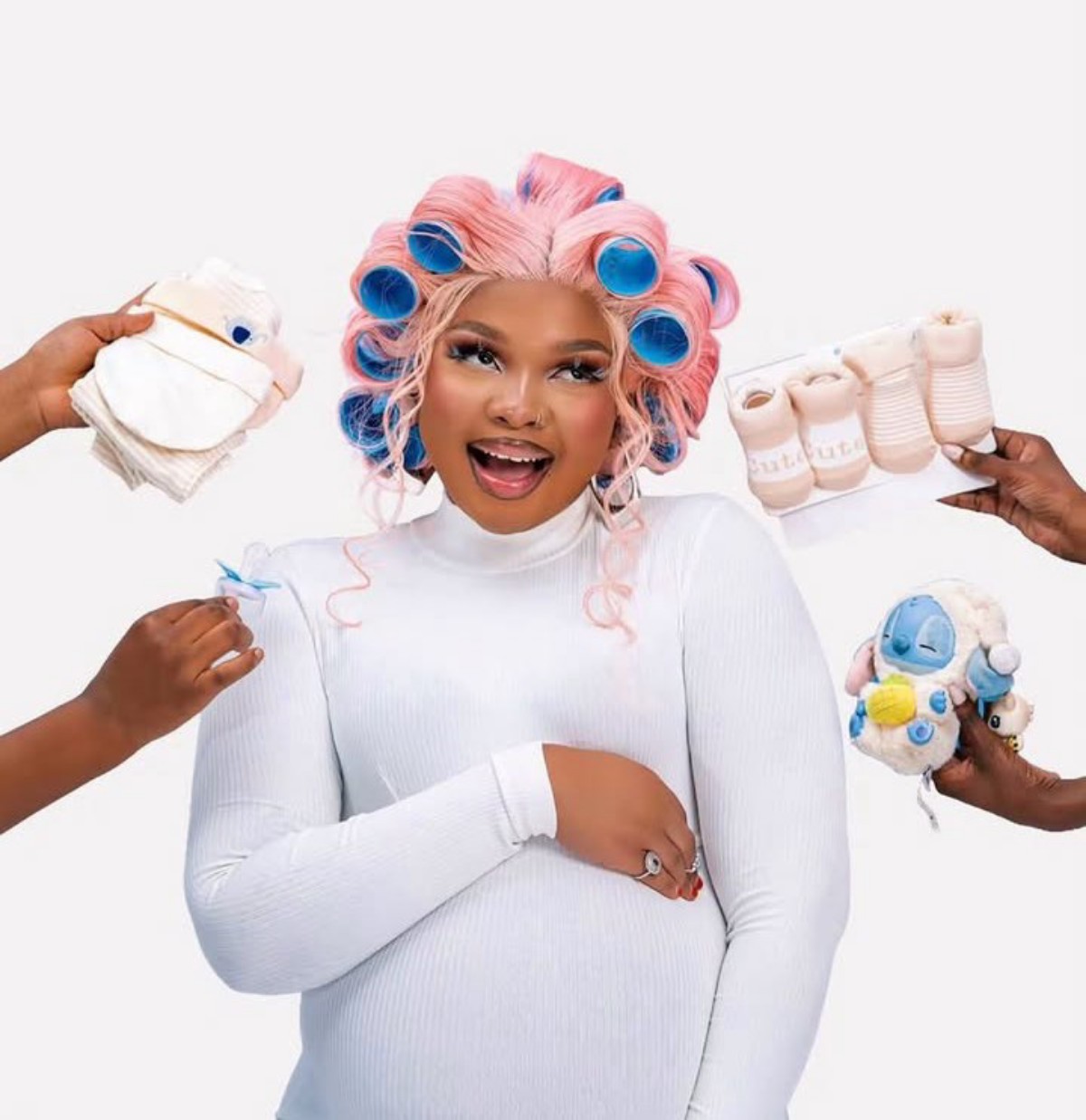 Pregnant woman smiling with pink-blue hair rollers, surrounded by hands offering baby items like wipes, socks, and plush toy.