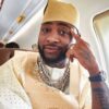 Portrait of a man in cream traditional attire and kufi hat, wearing multiple chains and a watch, seated inside an airplane.