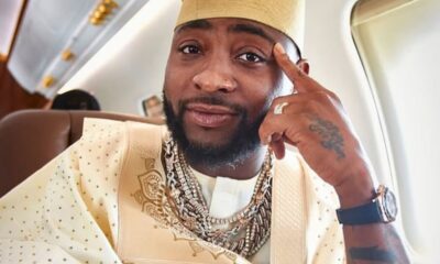 Portrait of a man in cream traditional attire and kufi hat, wearing multiple chains and a watch, seated inside an airplane.