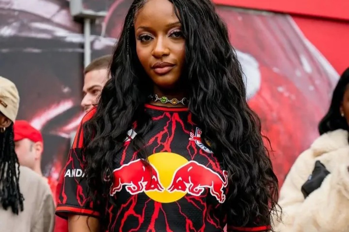 Portrait of a young woman with long wavy black hair wearing a Red Bull jersey with red lightning pattern, outdoors in a crowd experience set.