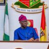 Man in blue traditional attire and purple cap seated at a podium with Nigerian flags behind him at a formal government setting.