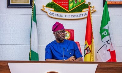 Man in blue traditional attire and purple cap seated at a podium with Nigerian flags behind him at a formal government setting.