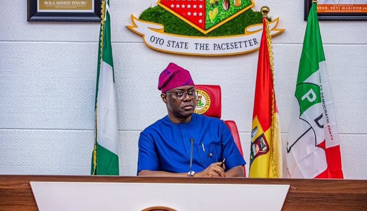 Man in blue traditional attire and purple cap seated at a podium with Nigerian flags behind him at a formal government setting.