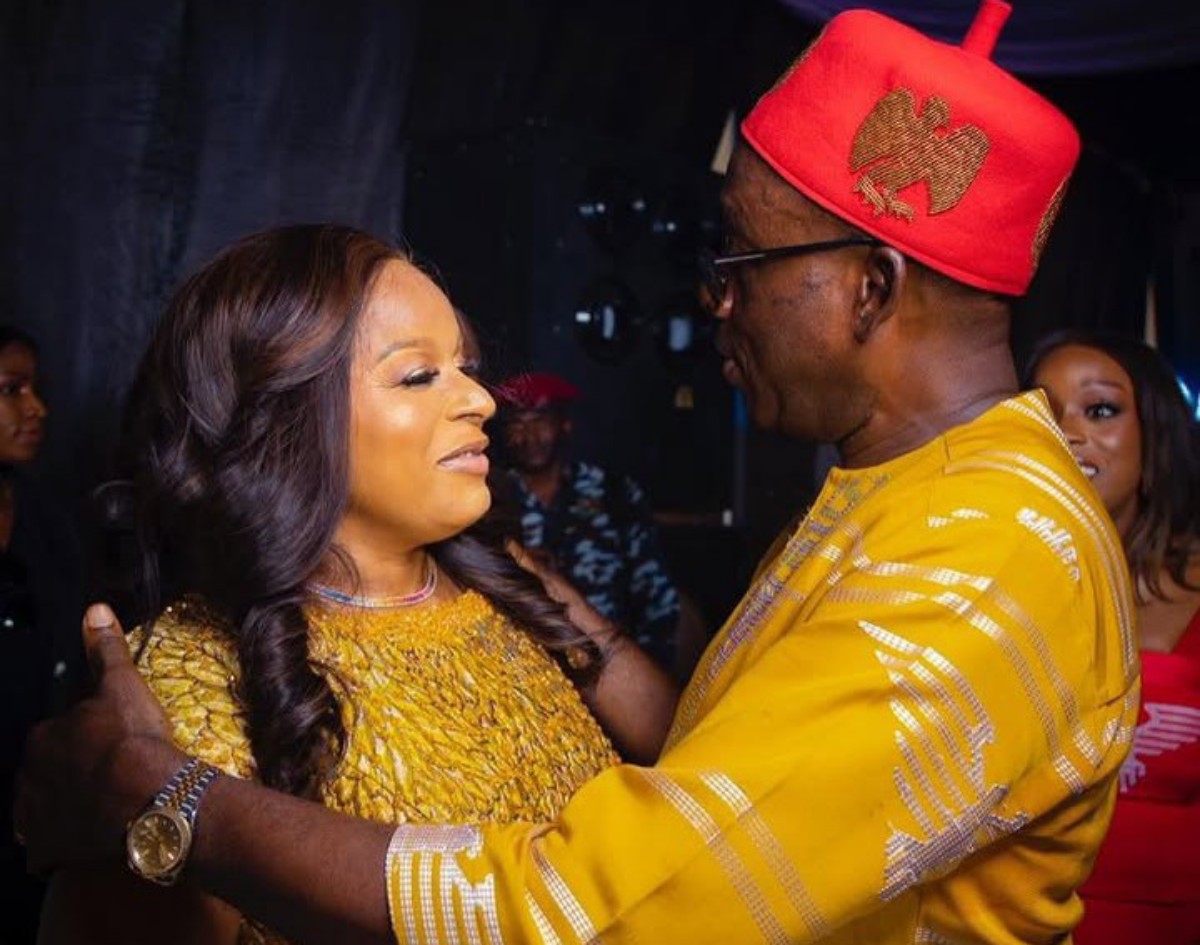 Couple dancing closely at a celebration: woman in a gold lace dress facing a man in a bright yellow traditional outfit and red hat.