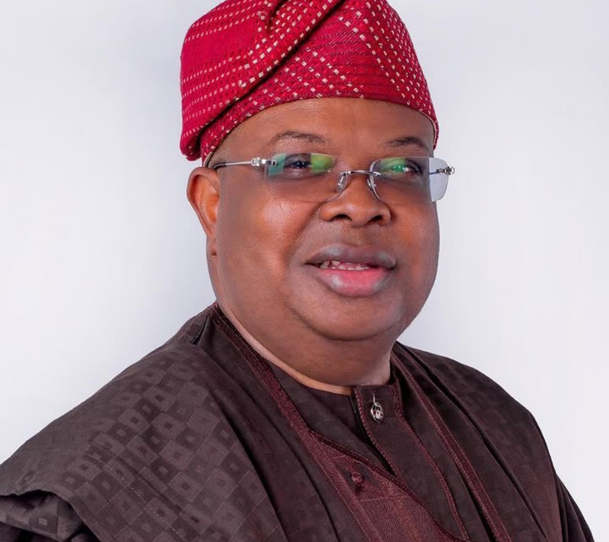 Smiling man in a brown traditional outfit with a red patterned cap and glasses, head-and-shoulders portrait.