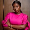 Confident Black woman wearing a bright pink blouse with tied sleeves, arms crossed, looking at the camera with a neutral expression, wearing jewelry.