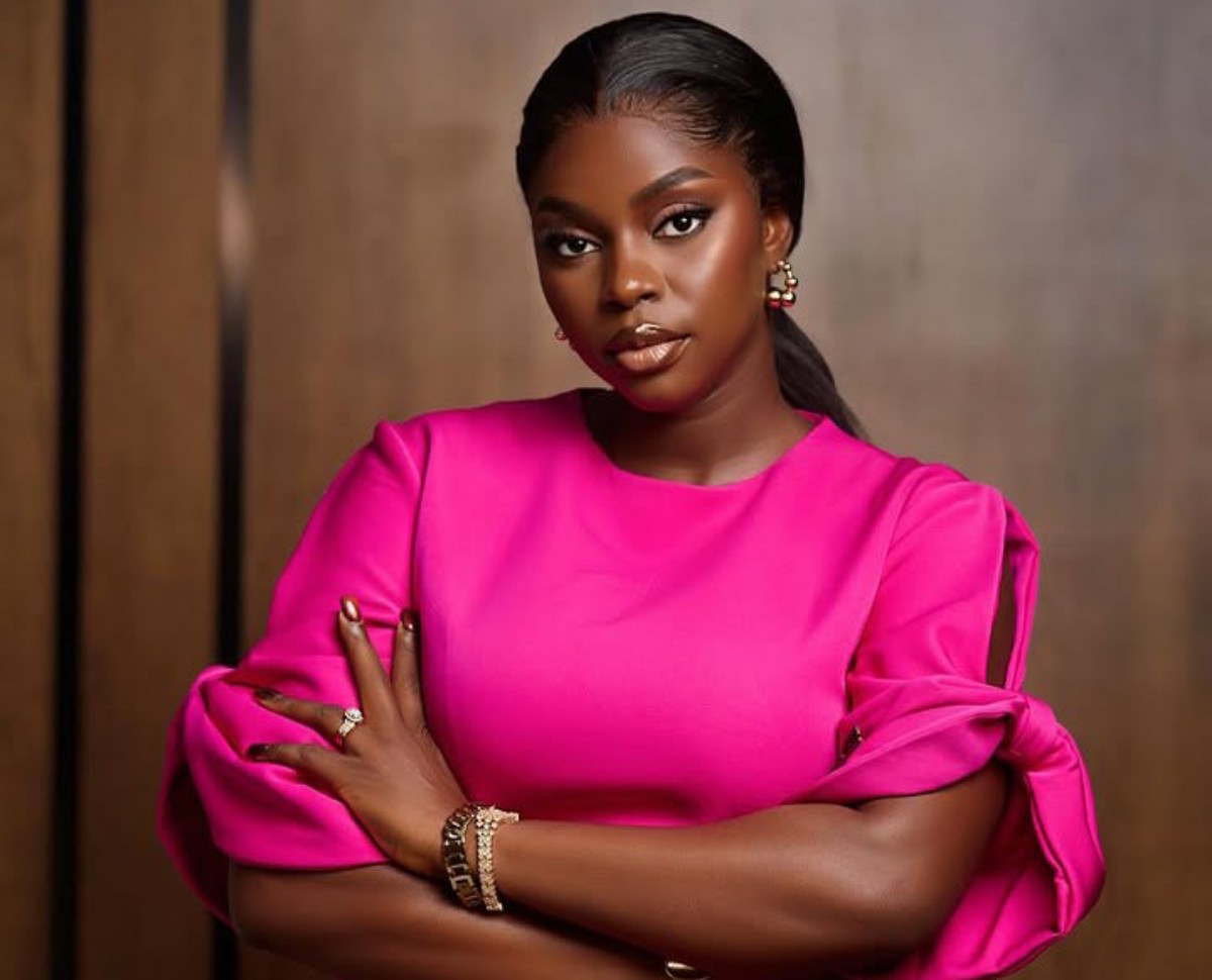 Confident Black woman wearing a bright pink blouse with tied sleeves, arms crossed, looking at the camera with a neutral expression, wearing jewelry.