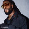 Man with a full beard and dreadlocks wearing amber-tinted sunglasses, a black shirt, a gold chain, and a textured black clutch resting on his arm.