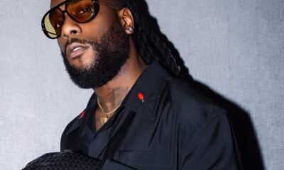 Man with a full beard and dreadlocks wearing amber-tinted sunglasses, a black shirt, a gold chain, and a textured black clutch resting on his arm.